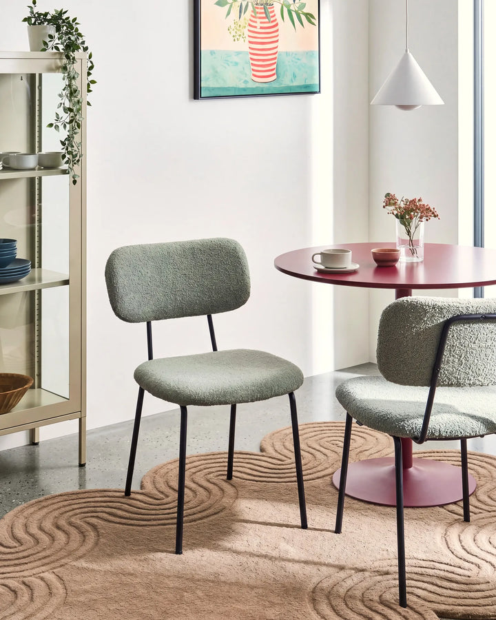 Light green bouclé dining chairs with curved backrests and black metal legs arranged around a dining table in a bright, modern dining space with minimalist décor.