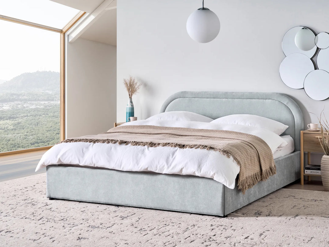 Gray upholstered bed with white bedding in a modern bedroom with large windows.