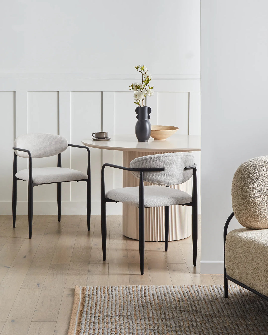 Off-white upholstered dining chairs gathered around a round pedestal table in a bright, minimalist dining space with light wood flooring, neutral décor, sculptural accents, and soft natural light.