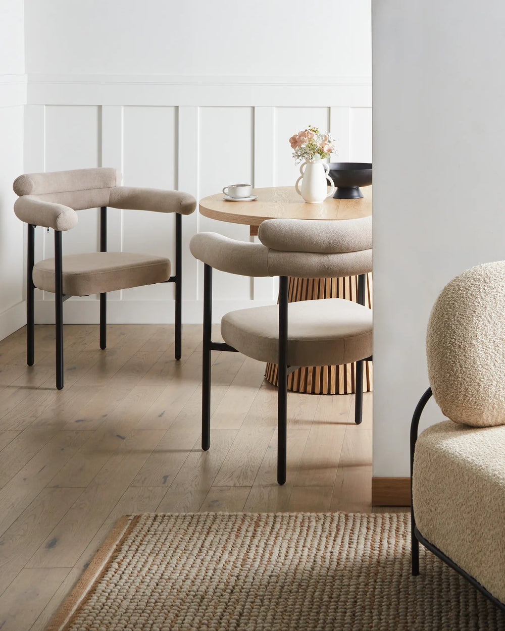 Beige fabric dining chairs with rounded backrests and black metal legs arranged around a dining table in a bright, modern dining space with minimalist décor.