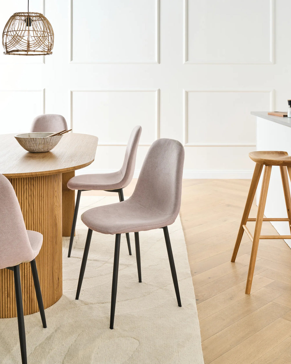 Beige dining chairs arranged around a round wood-top pedestal table in a bright, modern dining room with warm sunlight, a woven rug, minimalist décor, and soft neutral accents.