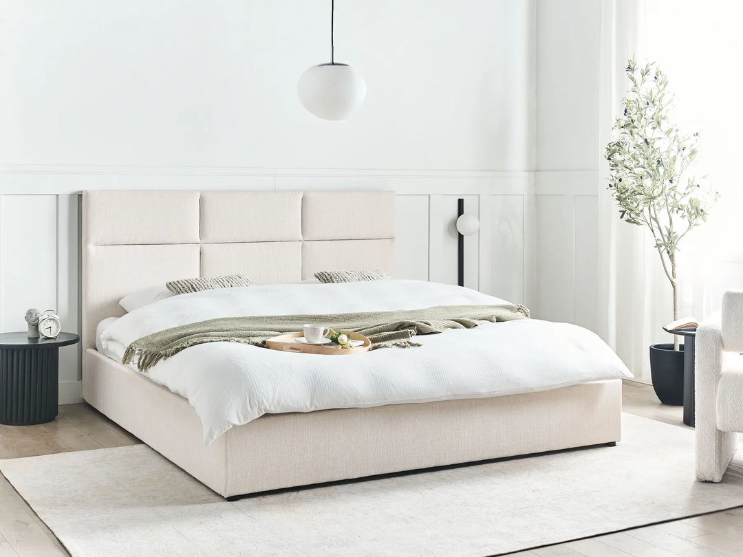 Beige upholstered bed in a minimalistic bedroom with white walls and light wood flooring.