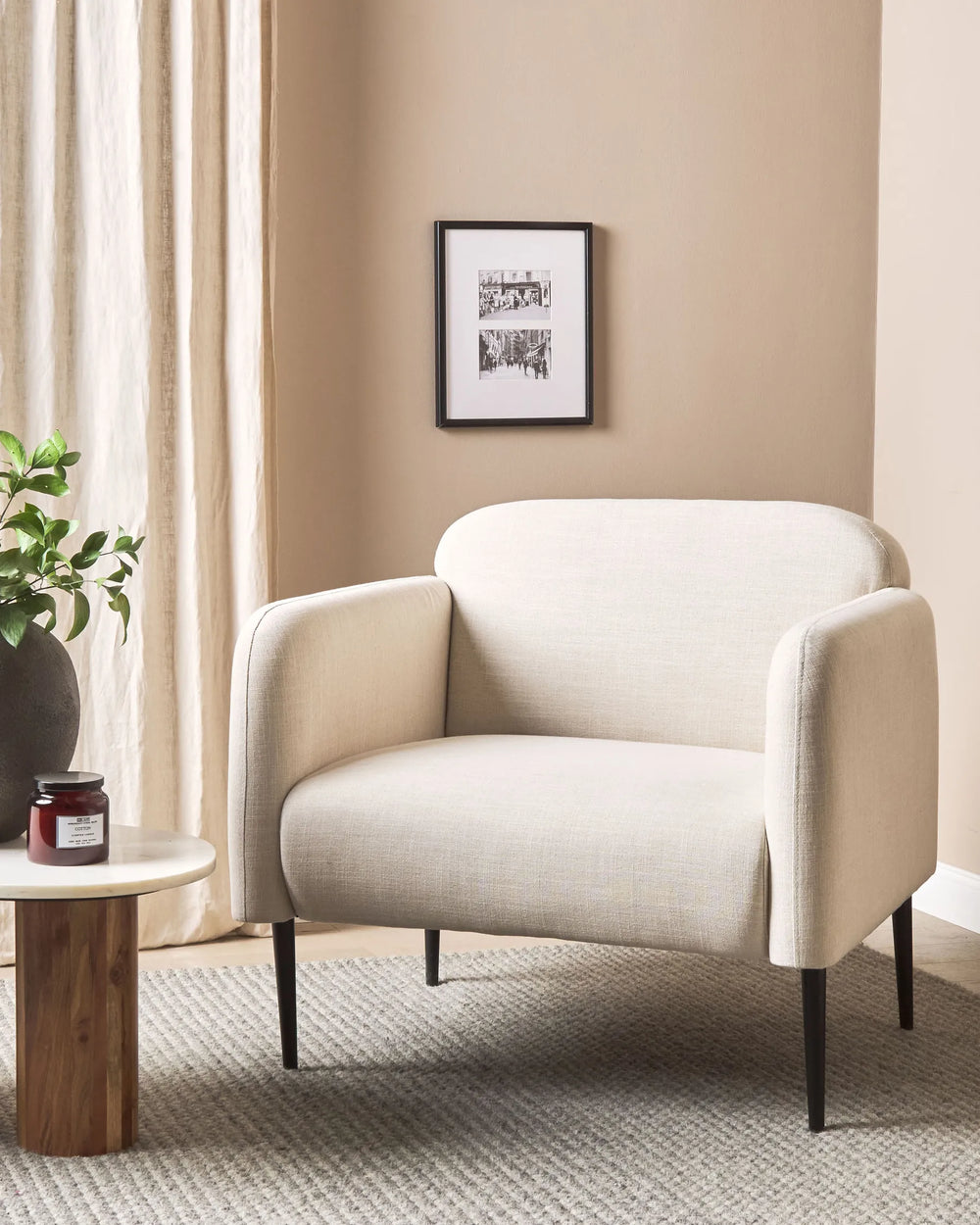 Beige armchair in a living room with a framed picture on the wall and a small table with a plant.