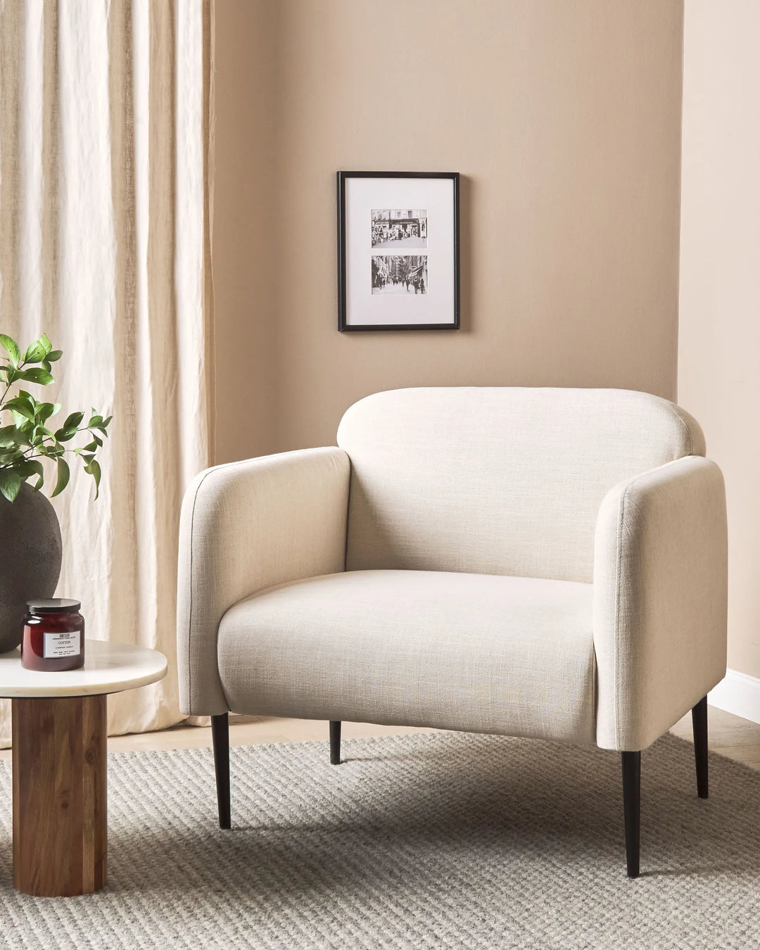 Beige armchair in a living room with a framed picture on the wall and a small table with a plant.