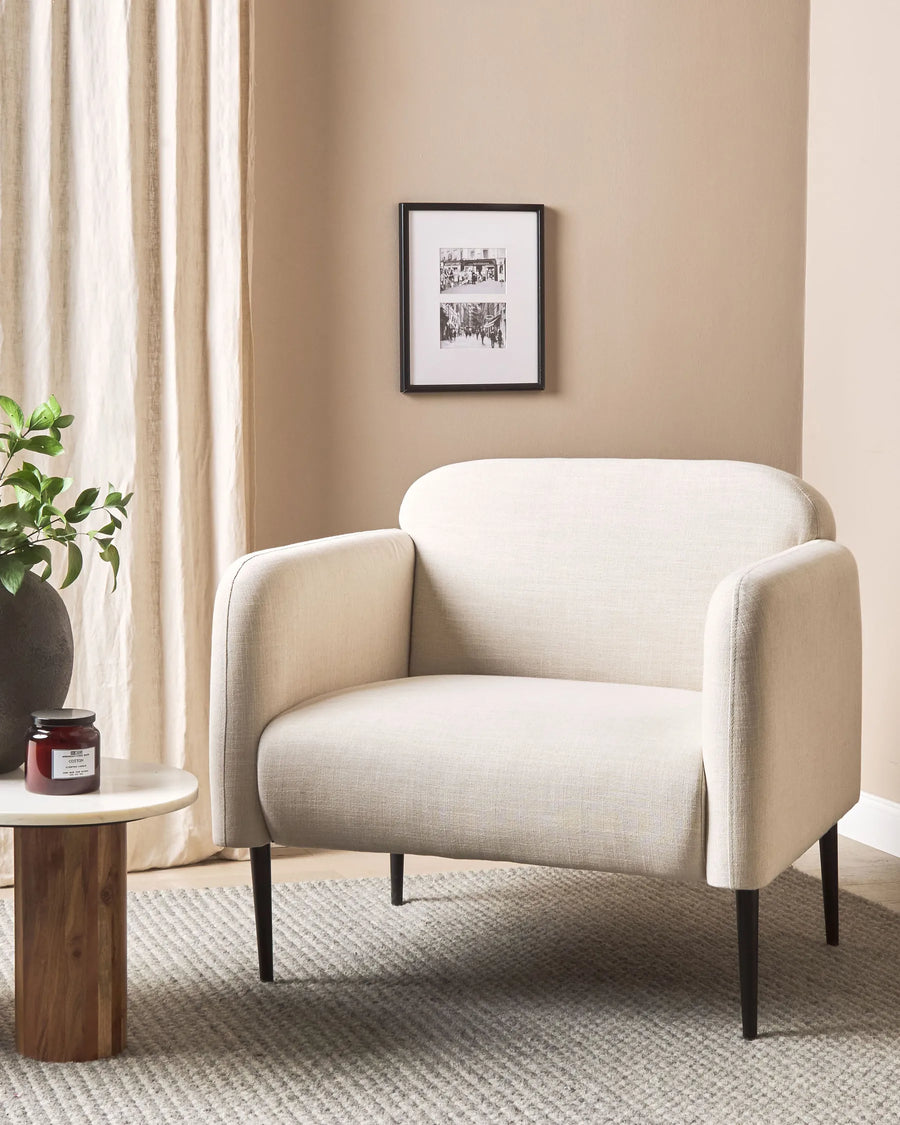 Beige armchair in a living room with a framed picture on the wall and a small table with a plant.