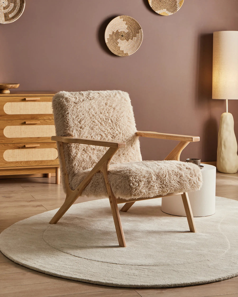Beige armchair with wooden frame in a room with a wooden dresser and lamp.