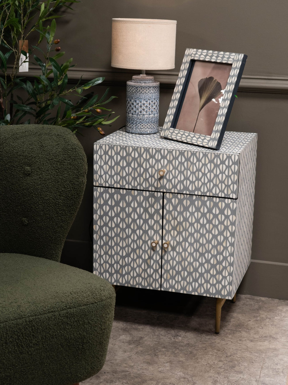 Patterned nightstand with a lamp and photo frame next to a green armchair.