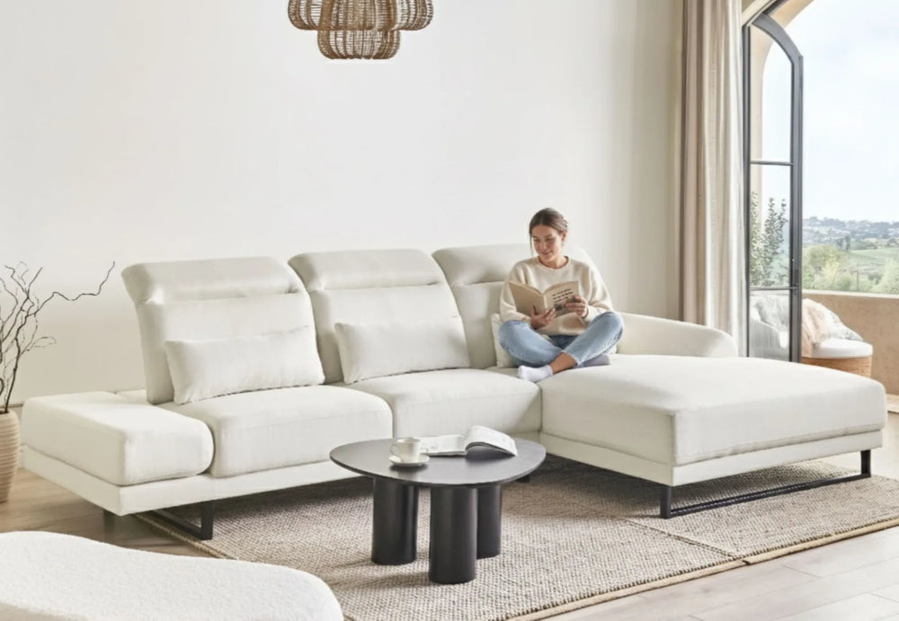 Woman reading a book on a white sectional sofa in a bright living room.