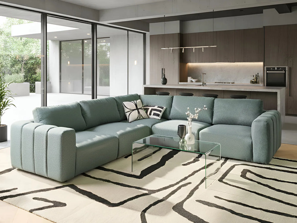 Modern living room with a light blue sectional sofa, glass coffee table, and kitchen in the background.