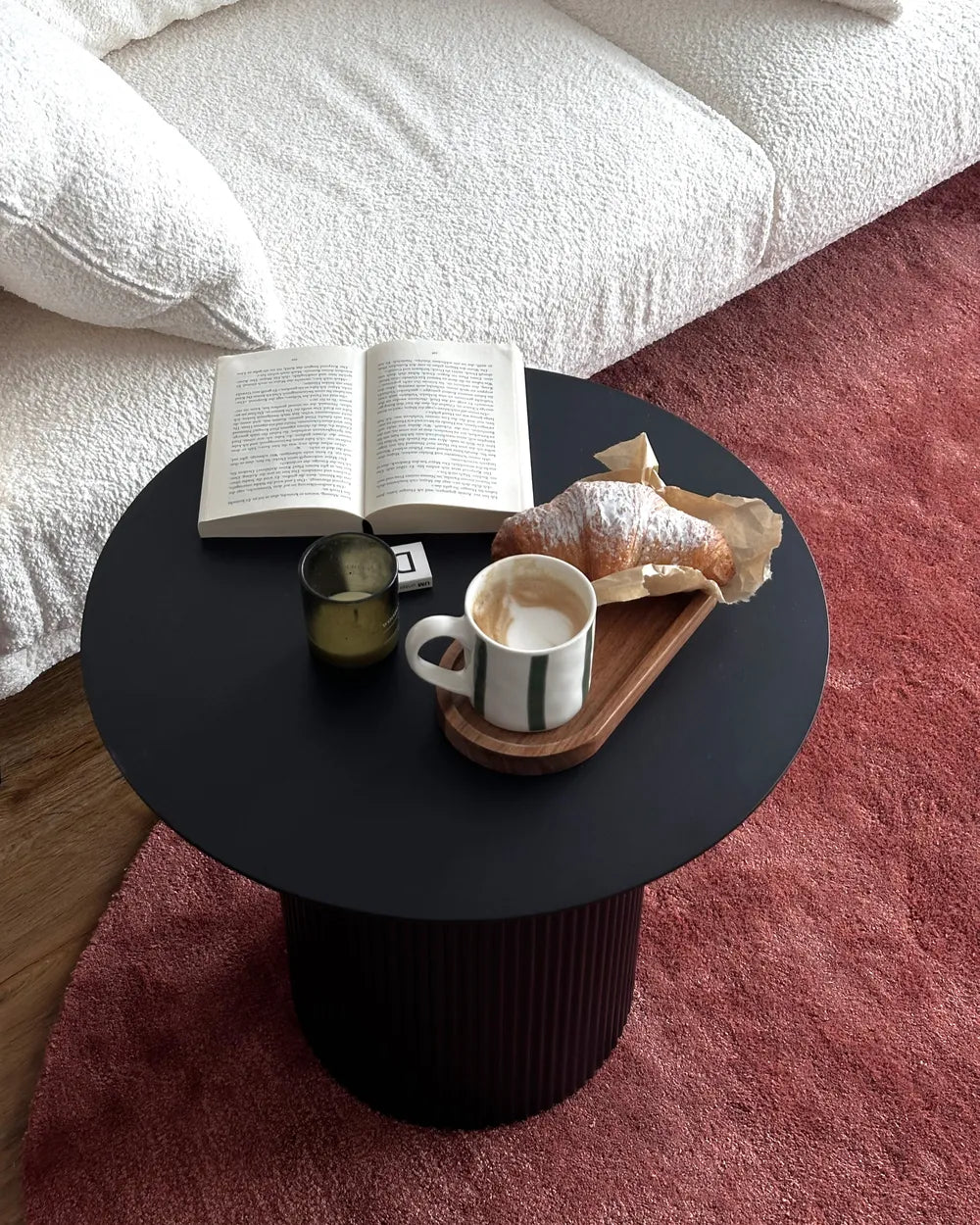 Black side table with a book, coffee cup, and pastry on a pink carpet.