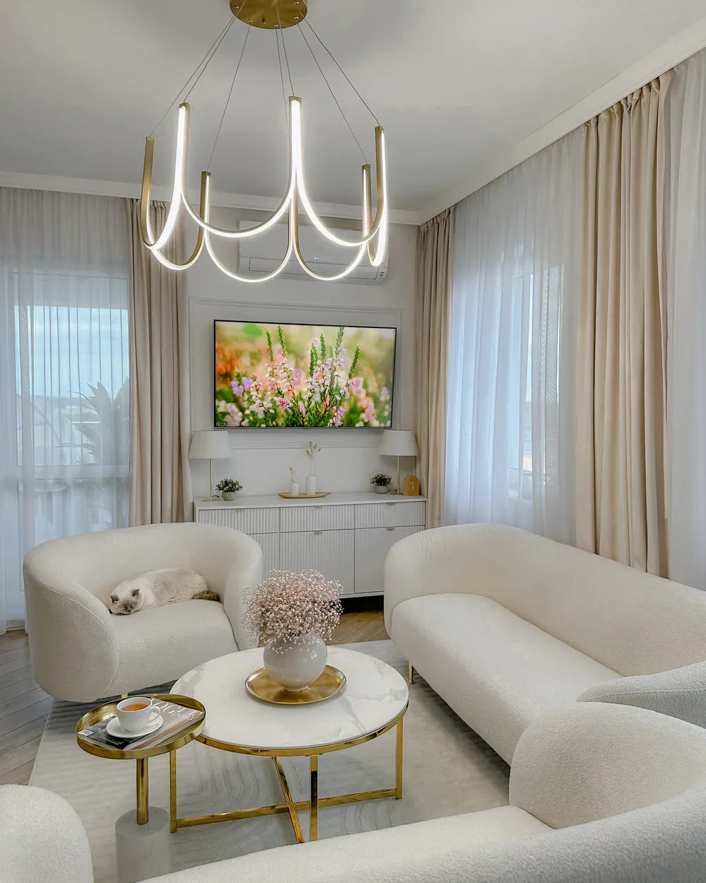 Modern living room with white furniture, a chandelier, and a TV displaying floral imagery.