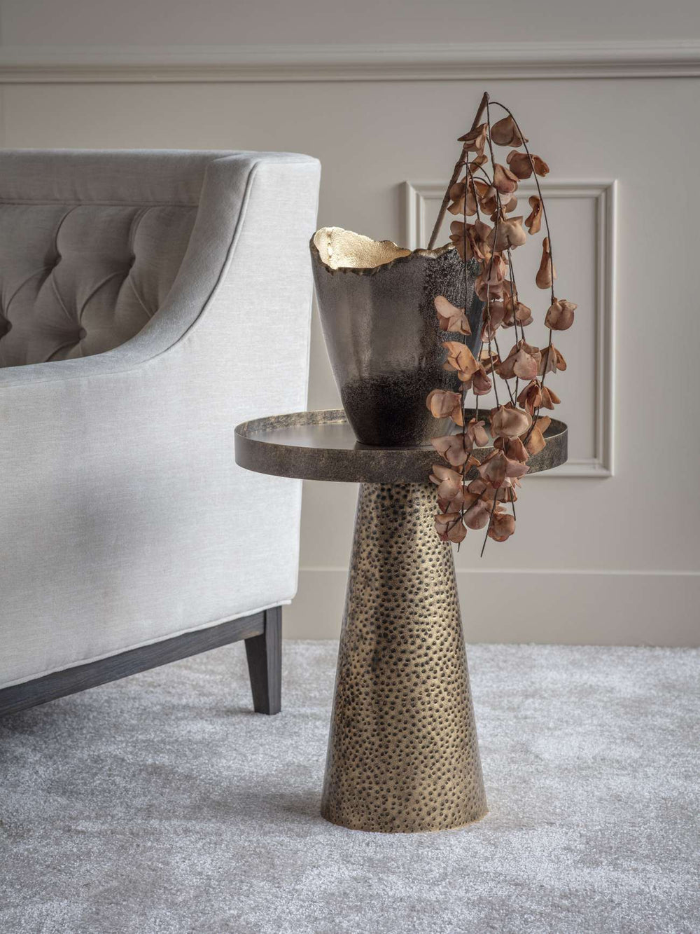 Decorative side table with a textured base and a vase with dried leaves in a living room setting.