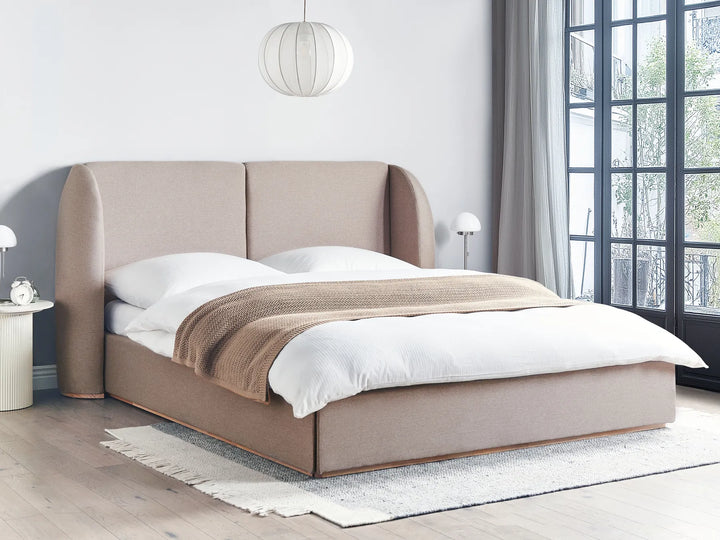 Beige upholstered bed in a modern bedroom with large windows.