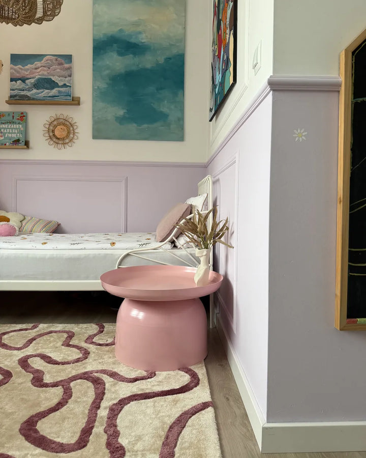 Pink stool in a room with a bed, artwork on the wall, and a patterned rug.