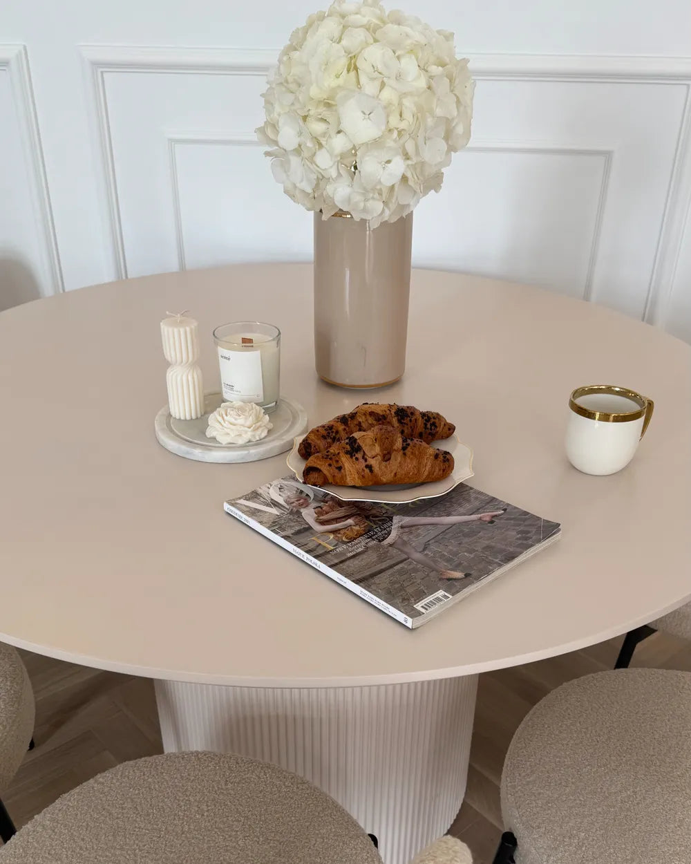 Round table with a vase of white flowers, croissants, candles, and a magazine on a light-colored floor.
