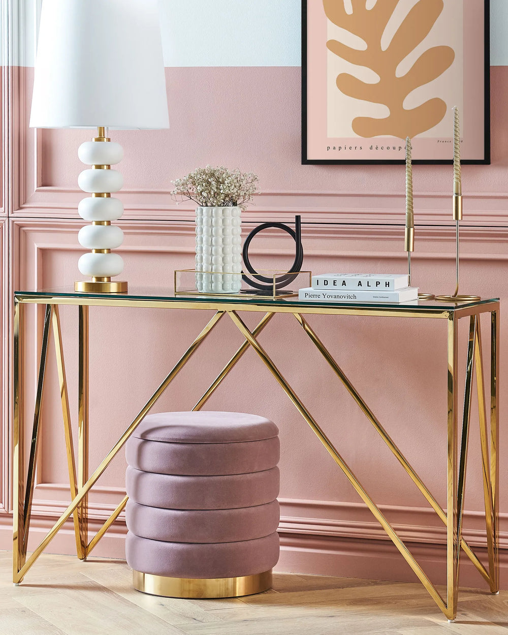Decorative console table with gold legs and a glass top, featuring a lamp, vase, and books against a pink wall.