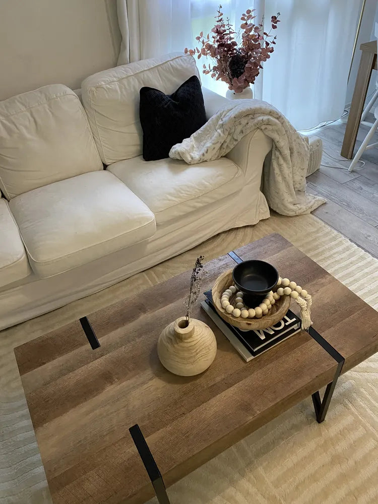 Beige sofa with a black pillow and white blanket, wooden coffee table with decorative items in a living room.