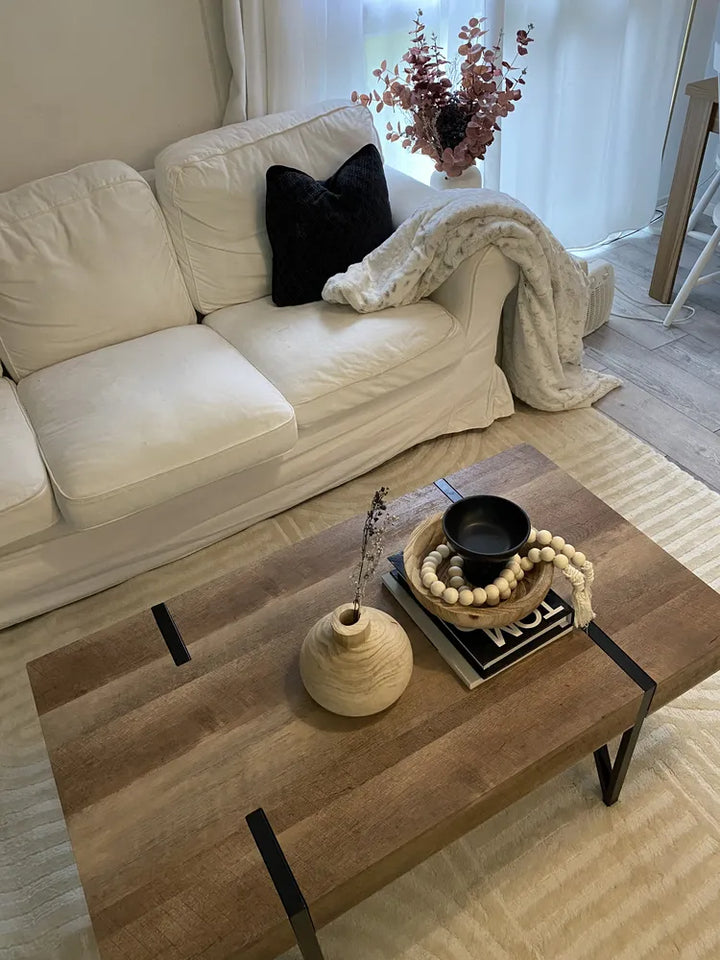 Beige sofa with a black pillow and white blanket, wooden coffee table with decorative items in a living room.