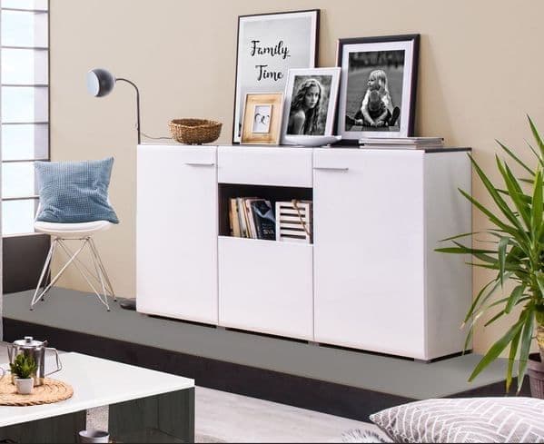 A modern white gloss sideboard with black accents, featuring drawers and doors, placed in a living room setting with decorative items and a framed photo on top.